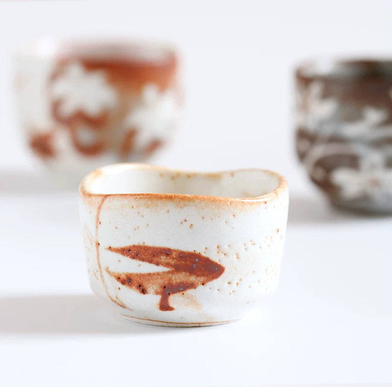 Close-up of a small, square-ish, white ceramic tea cup featuring a distinctive brown painted mark.