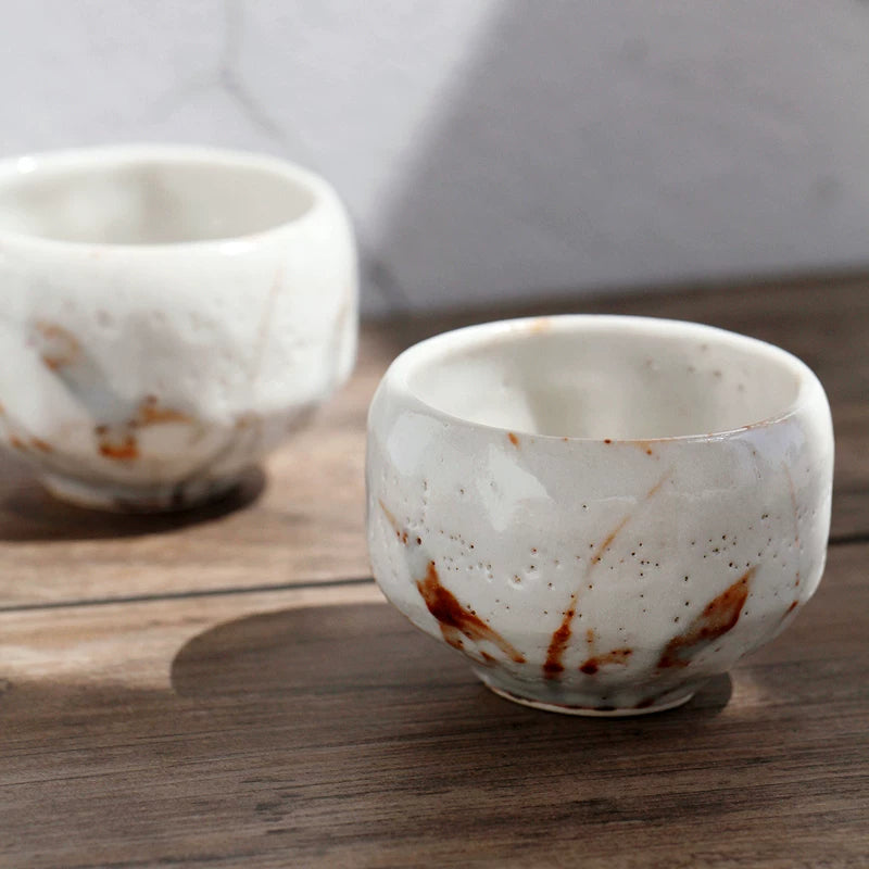 Two rustic ceramic tea cups, one in foreground, with white glaze and brown artistic streaks on a wooden surface.