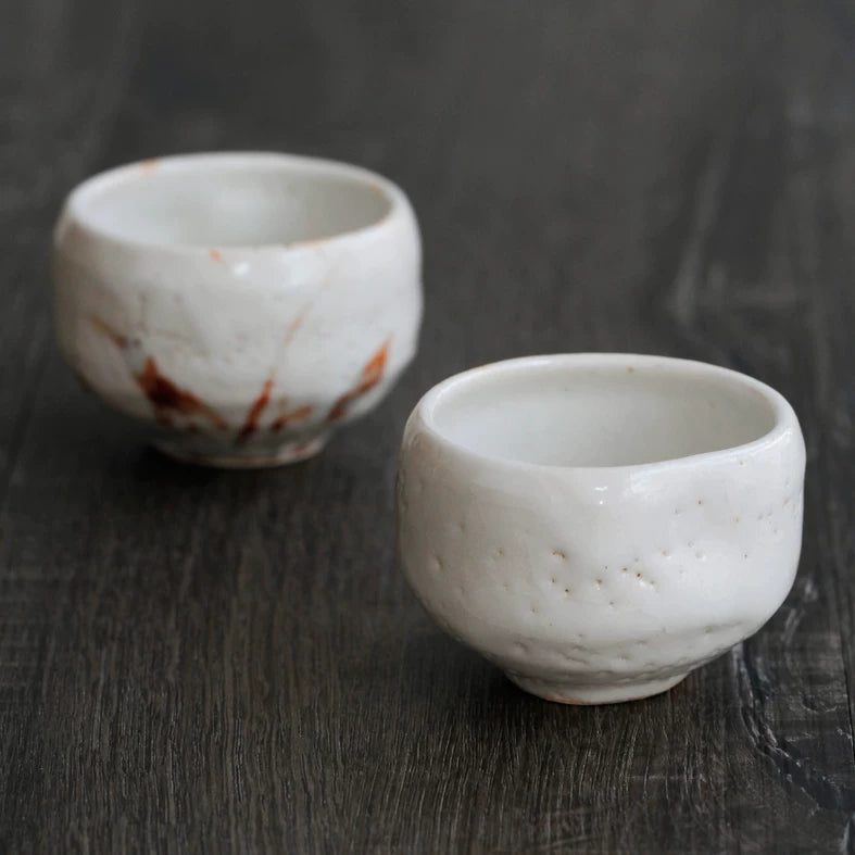 Two small, irregular, white ceramic tea cups with faint brown markings on a dark wooden table.