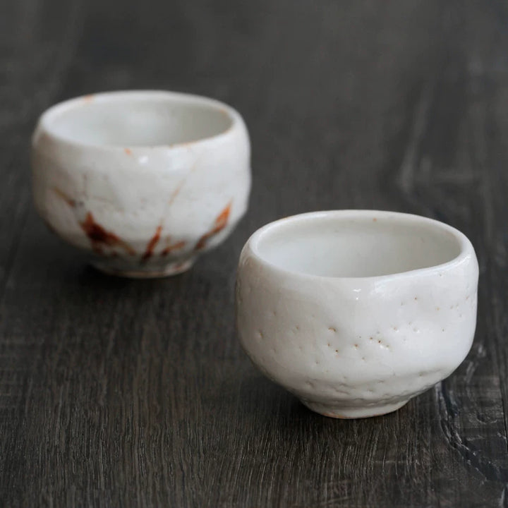 Two small, irregular, white ceramic tea cups with faint brown markings on a dark wooden table.
