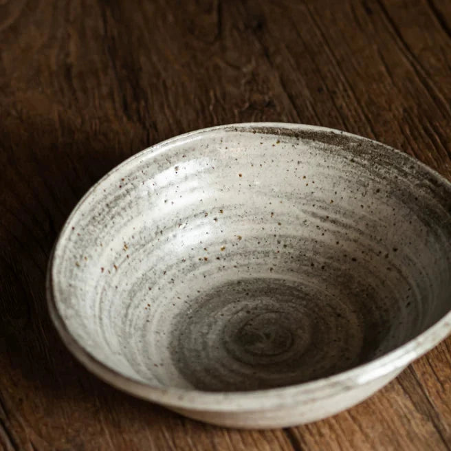 Close-up of a speckled cream and gray ceramic bowl with an irregular rim on a wooden surface.