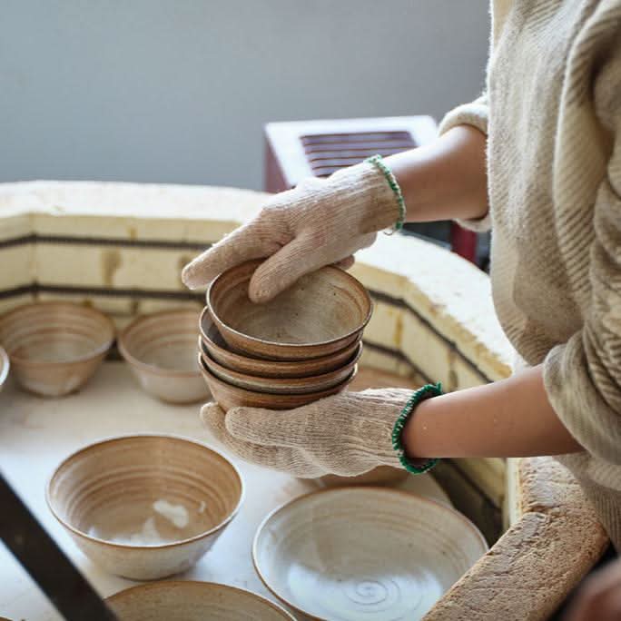 Hands in gloves hold a stack of brown clay bowls from a handmade tableware set.