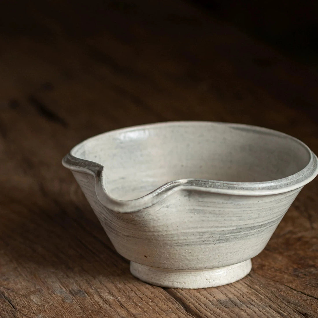 Side profile of a handmade gray and white ceramic tea bowl with an elegant pouring spout, on wood.