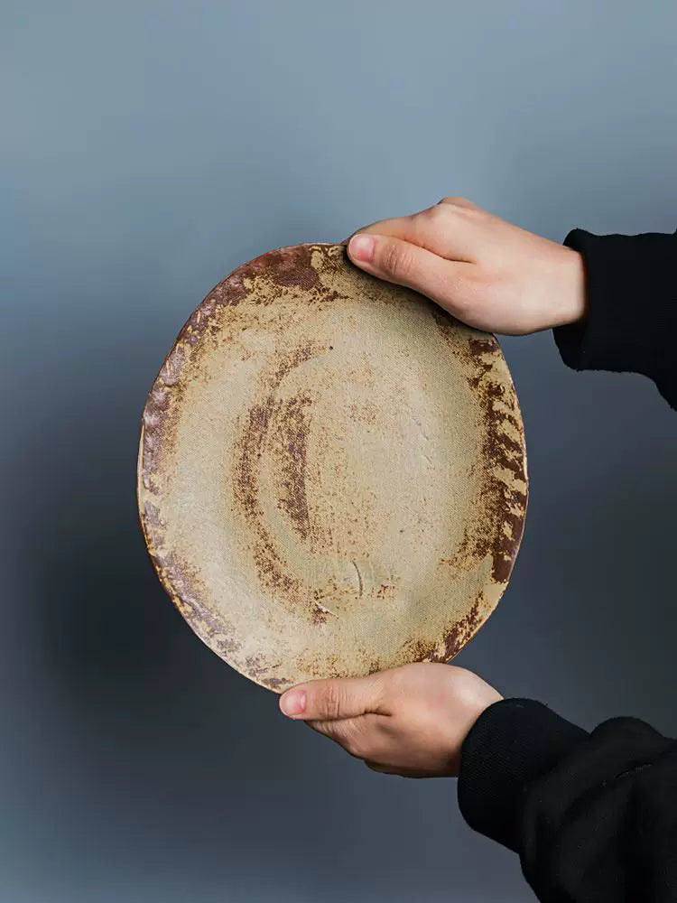 Hands presenting the back or underside of a textured, beige ceramic plate against a solid blue-gray wall.