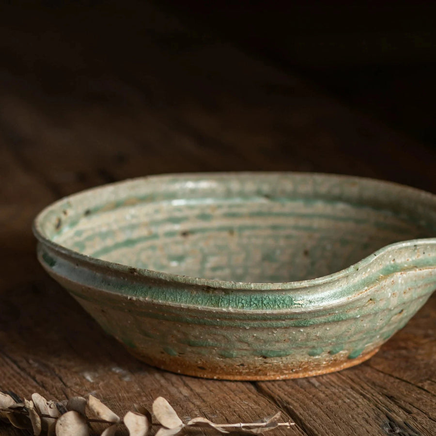 Close-up of a rustic, shallow ceramic bowl with a distinctive spout, featuring a mottled green glaze.