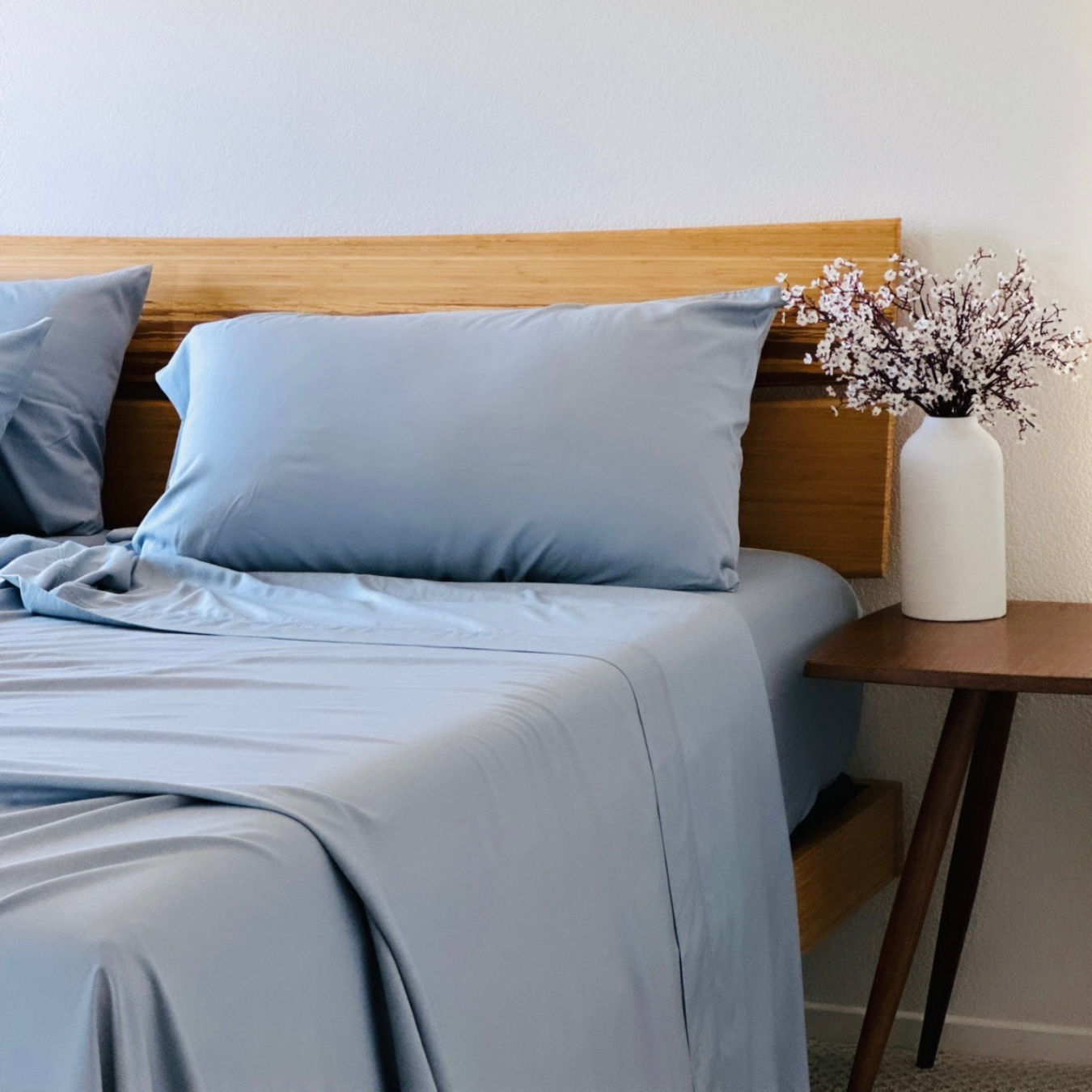 Light blue Copper Infused Bamboo Pillowcase set on a bed with wood headboard and white vase of flowers.