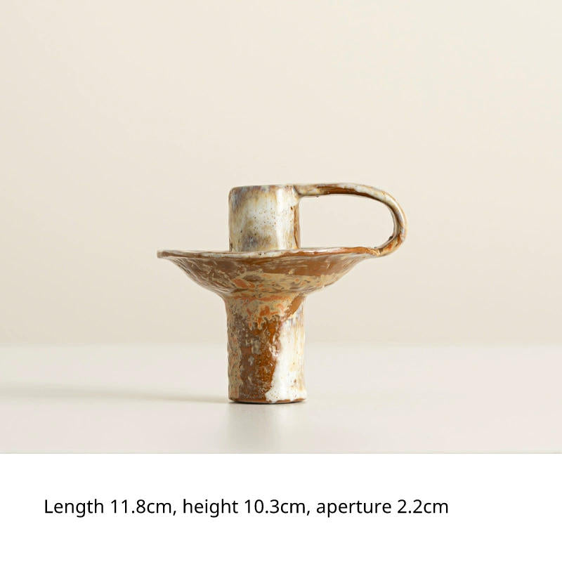 A rustic, hand-crafted ceramic candlestick with a unique, abstract design, featuring a flared base, a cylindrical stem, and a looped handle. The glaze has a variegated earthy brown and white mottled finish.