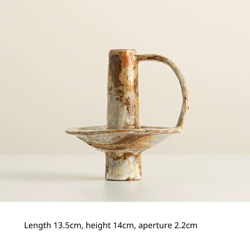 A rustic, handmade ceramic candlestick with a wide base and a curved handle, showcasing earthy tones of brown, beige, and white.