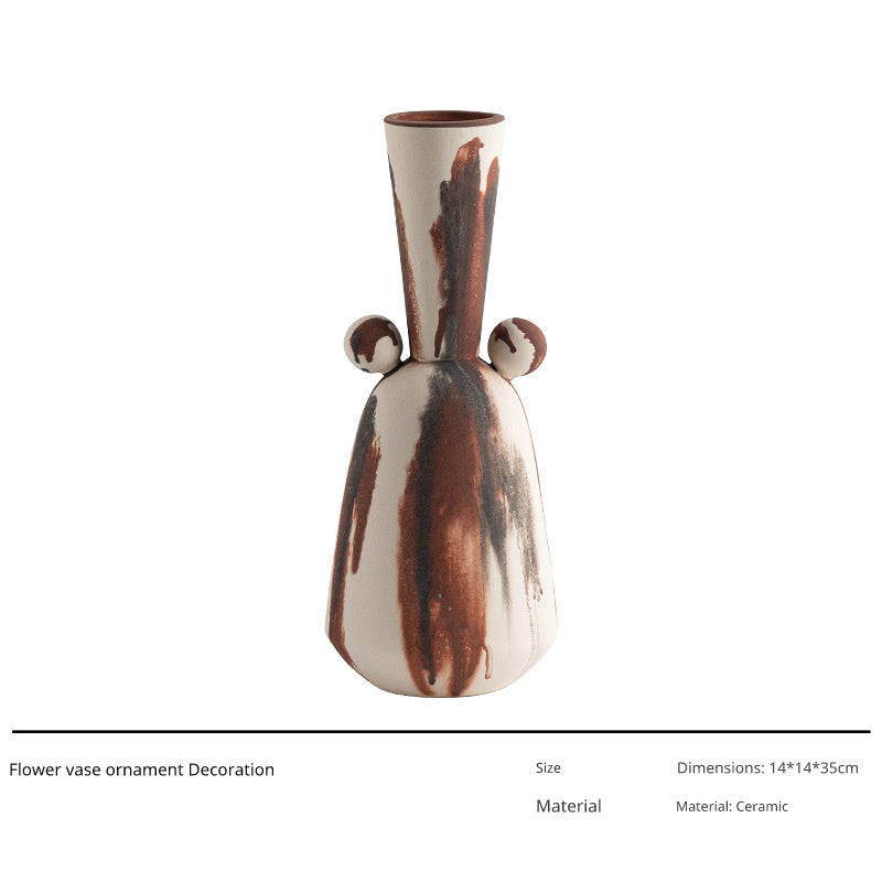 A unique ceramic vase with a white base color and abstract brown and black dripped patterns, featuring two small decorative spheres on either side of its slender neck.