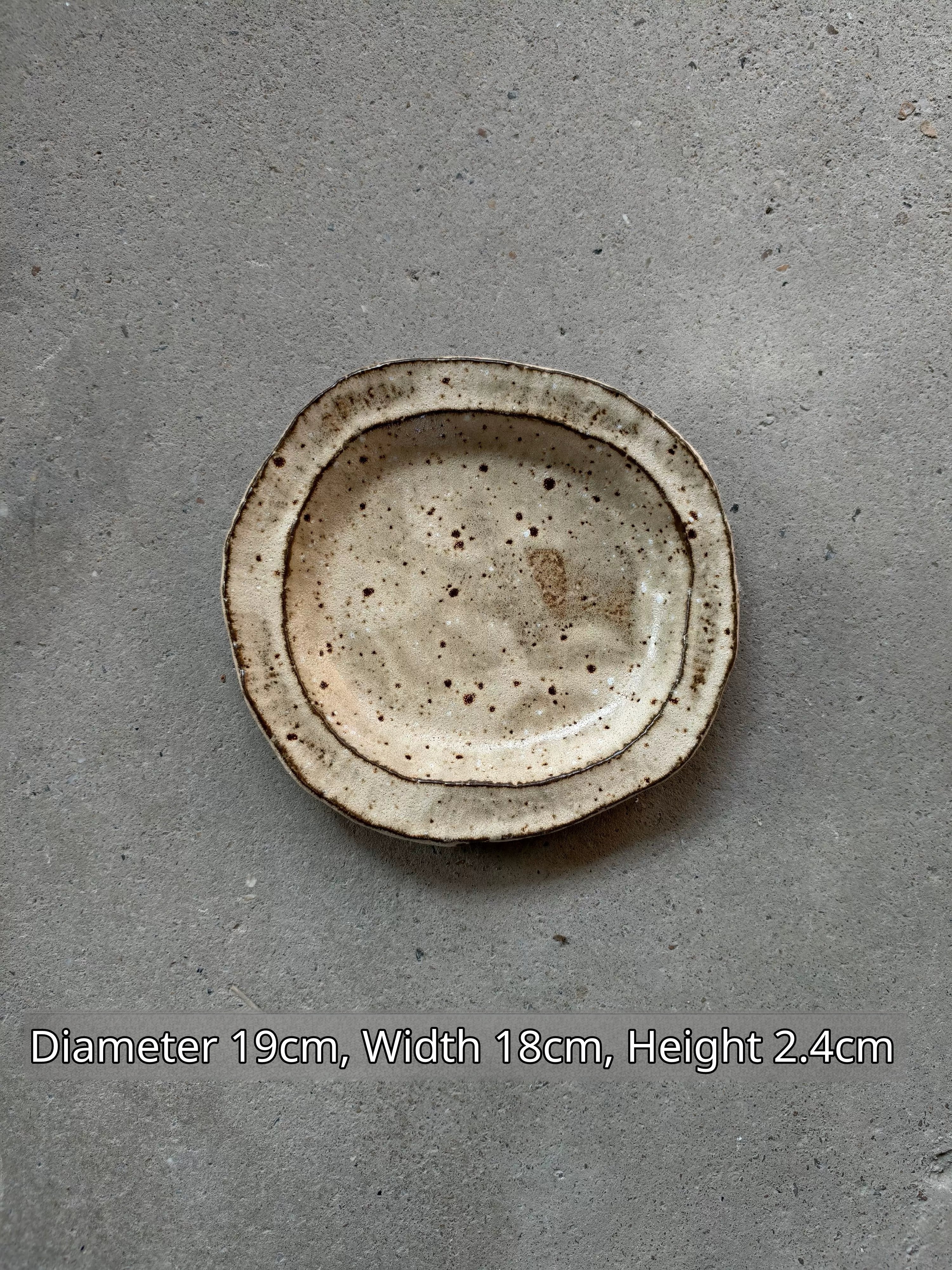 Oval ceramic plate in speckled beige and brown, 19x18x2.4cm, against grey concrete.