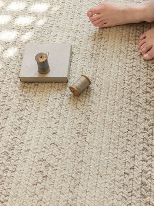 Natural fiber Round Wool Braided Rug in neutral tones, with spools of thread and a barefoot for scale.