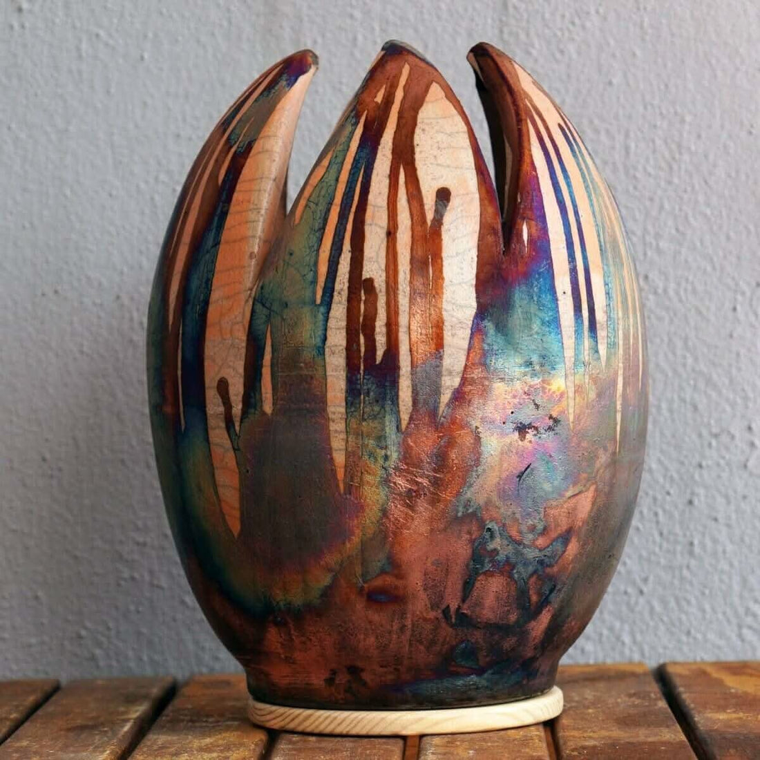 Large, tulip-shaped ceramic vase; raku-fired; colorful copper, bronze, and blue iridescence; wooden base.