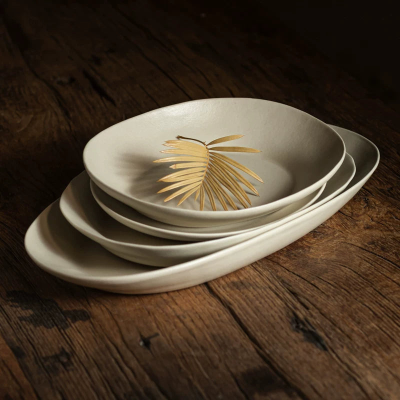 Stack of three oval, shallow ceramic serving dishes on dark wood, one topped with a dried palm leaf.