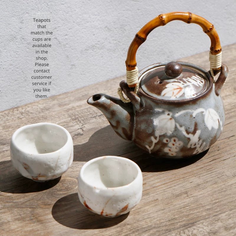 Small white ceramic tea cups next to a rustic, dark teapot with a bamboo handle on a wooden surface.