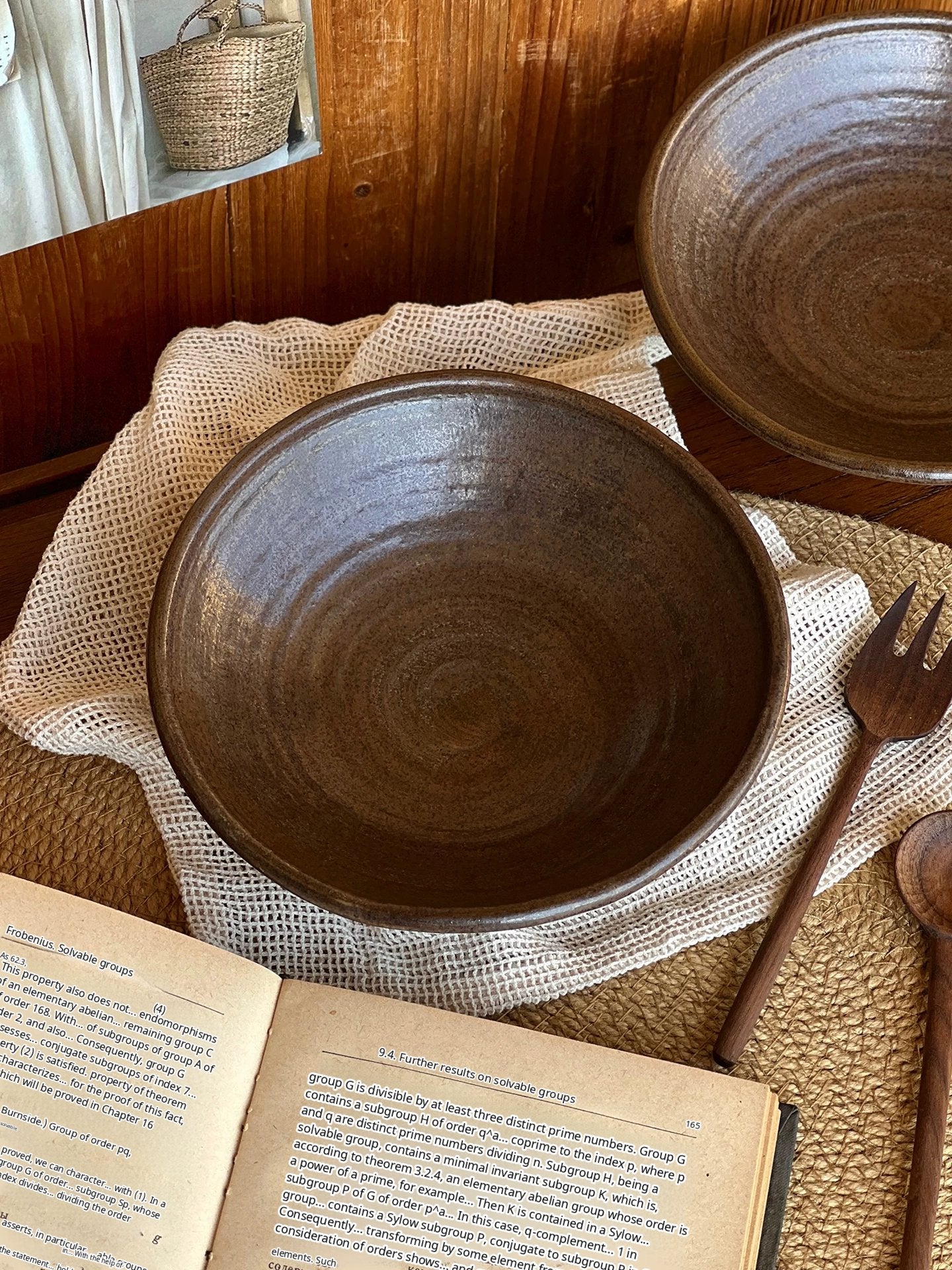A handcrafted dark brown ceramic bowl placed on a beige woven placemat next to an open book.
