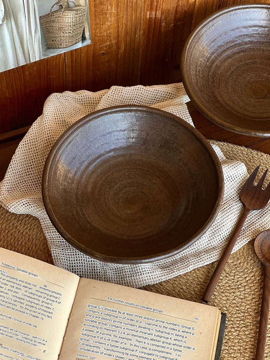 A handcrafted dark brown ceramic bowl placed on a beige woven placemat next to an open book.