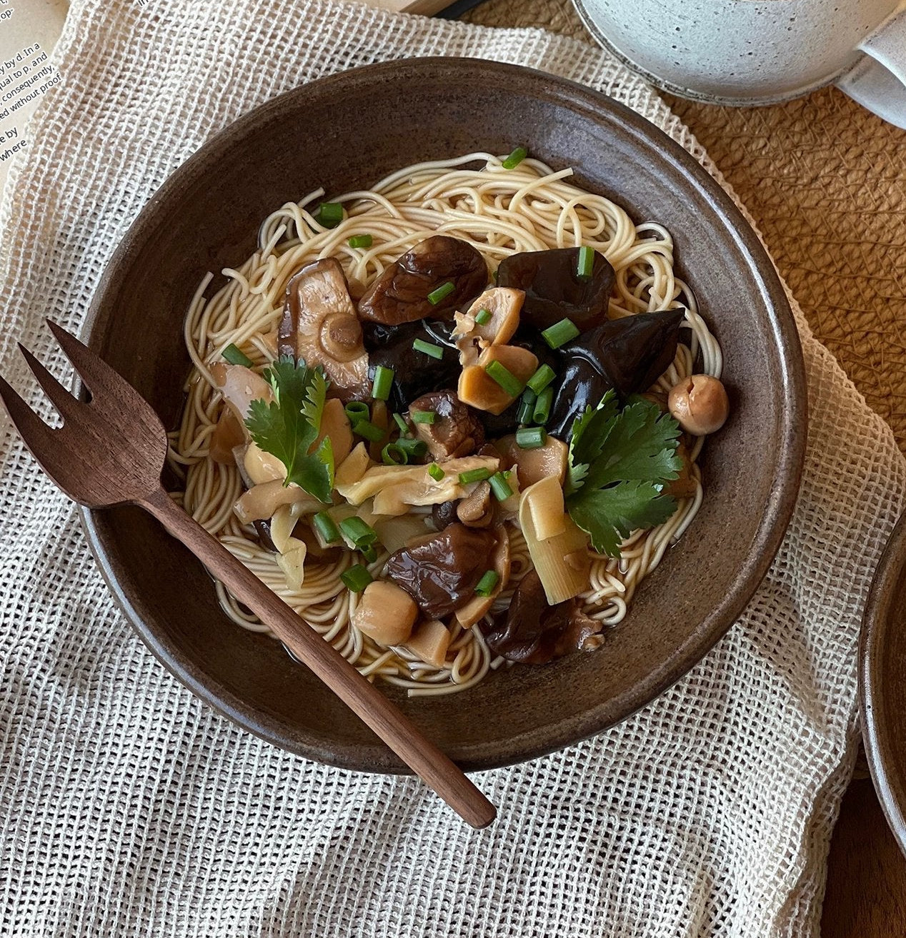 A ceramic bowl filled with noodles, mushrooms, and greens, served with a wooden fork and a cup of tea.