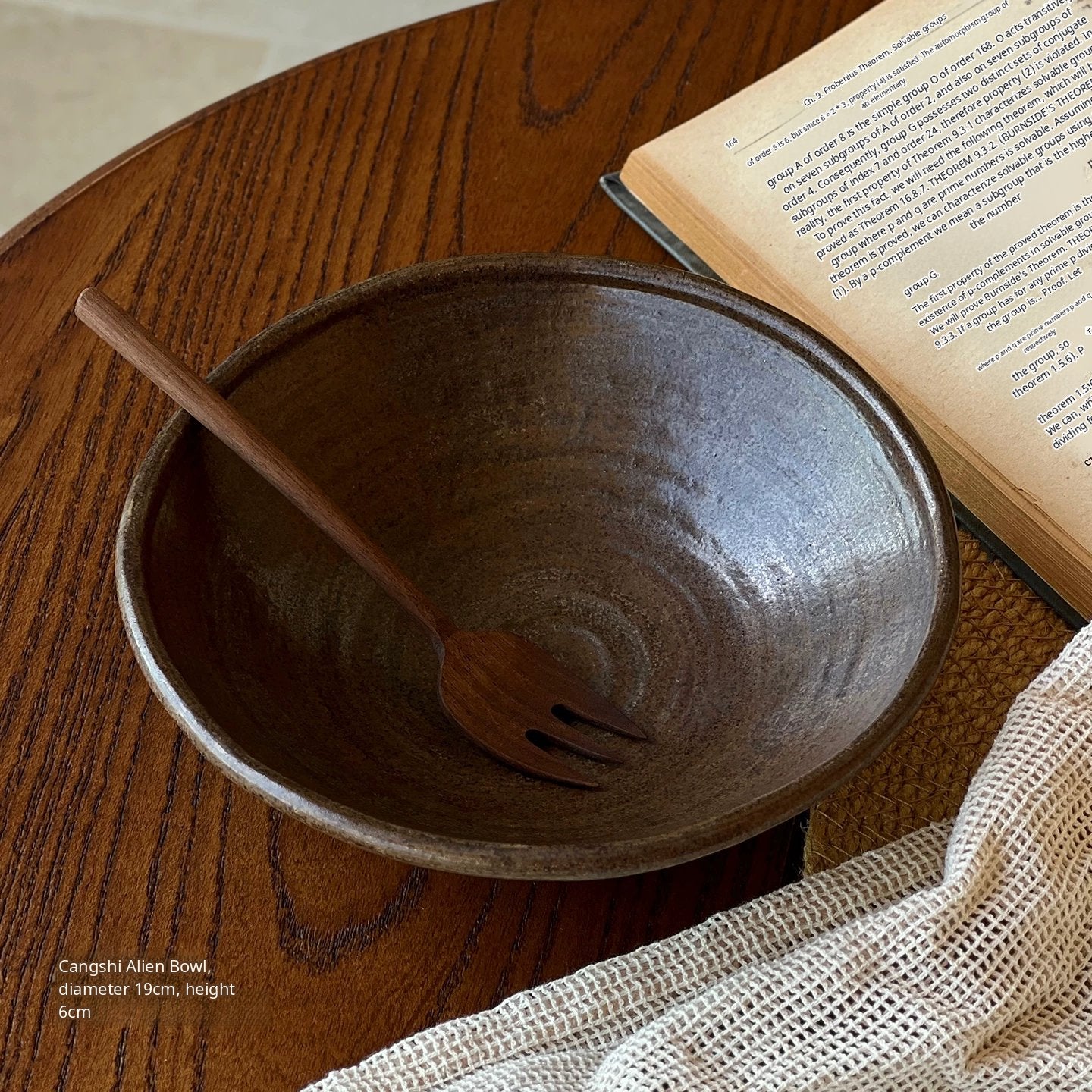 A deep dark brown ceramic bowl with a wooden fork, placed on a wooden table next to an open book.