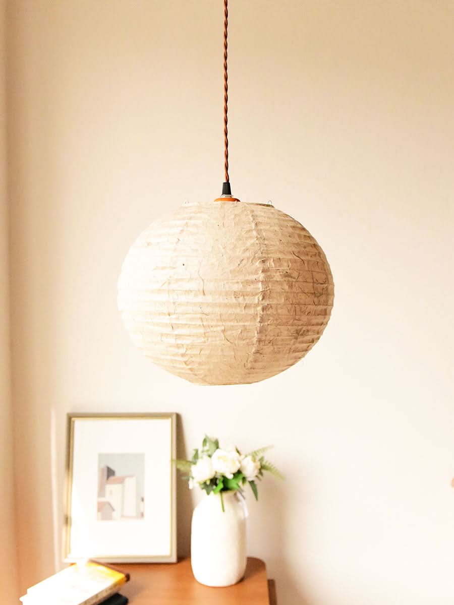 Textured, natural fiber handmade paper lampshade in off-white hangs over a wooden table with art and flowers.