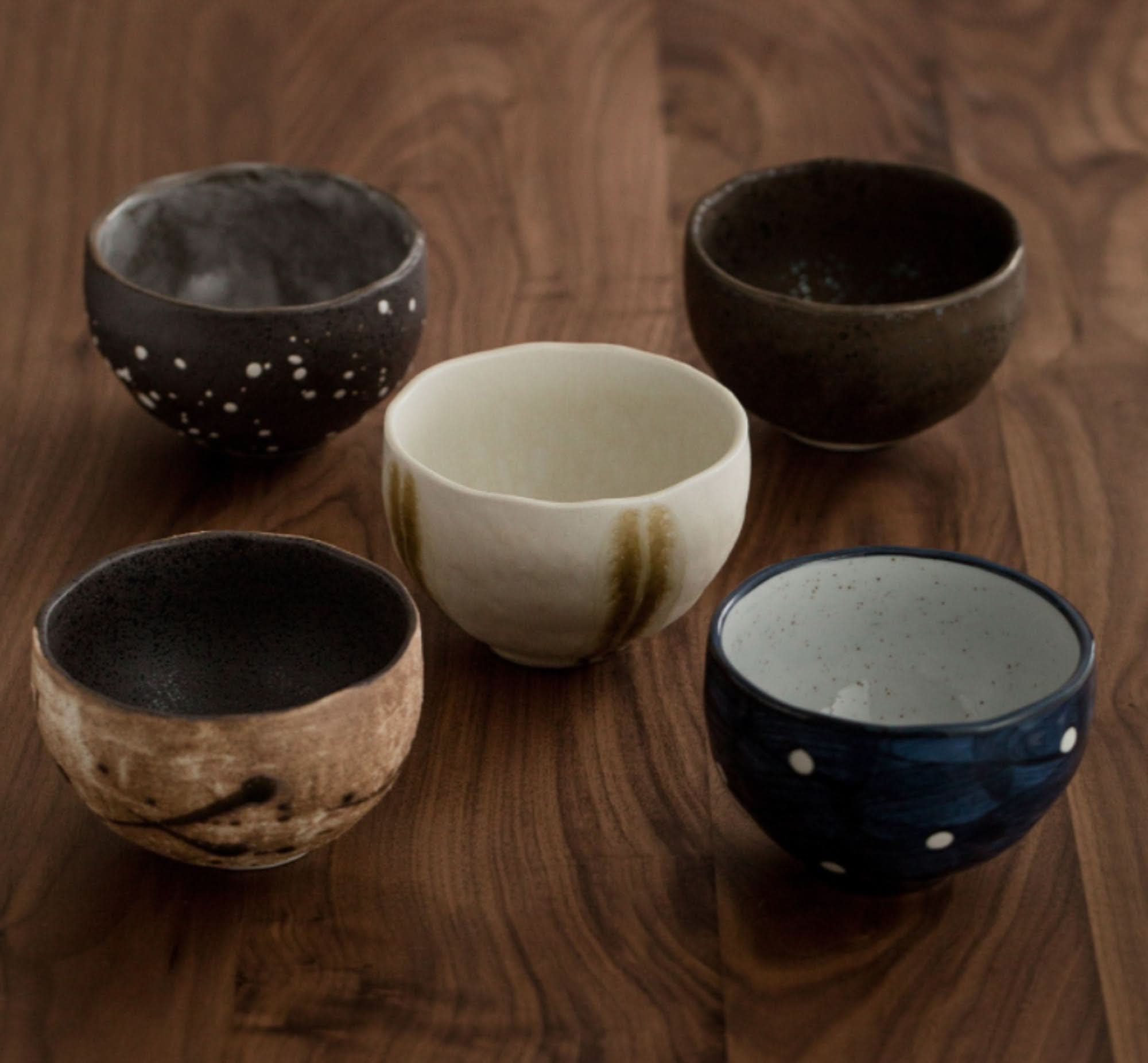 Assortment of five ceramic Japanese tea set cups in various earthy tones and patterns on a wooden surface.