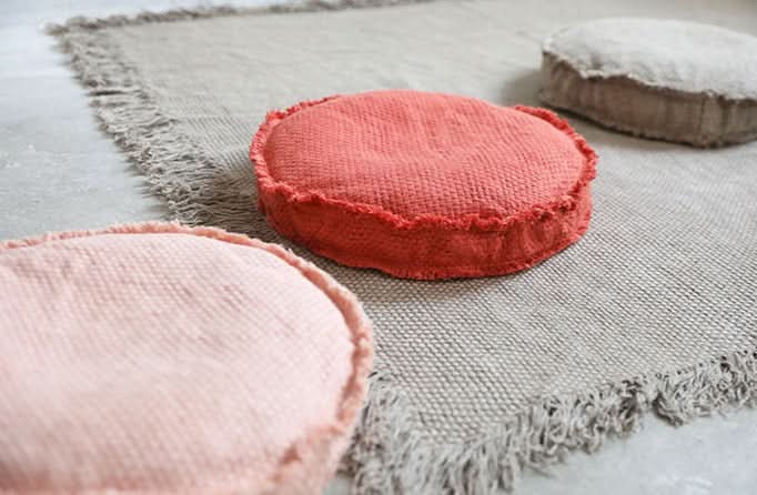 Round customizable textured floor cushion in coral and pink on a fringed rug.