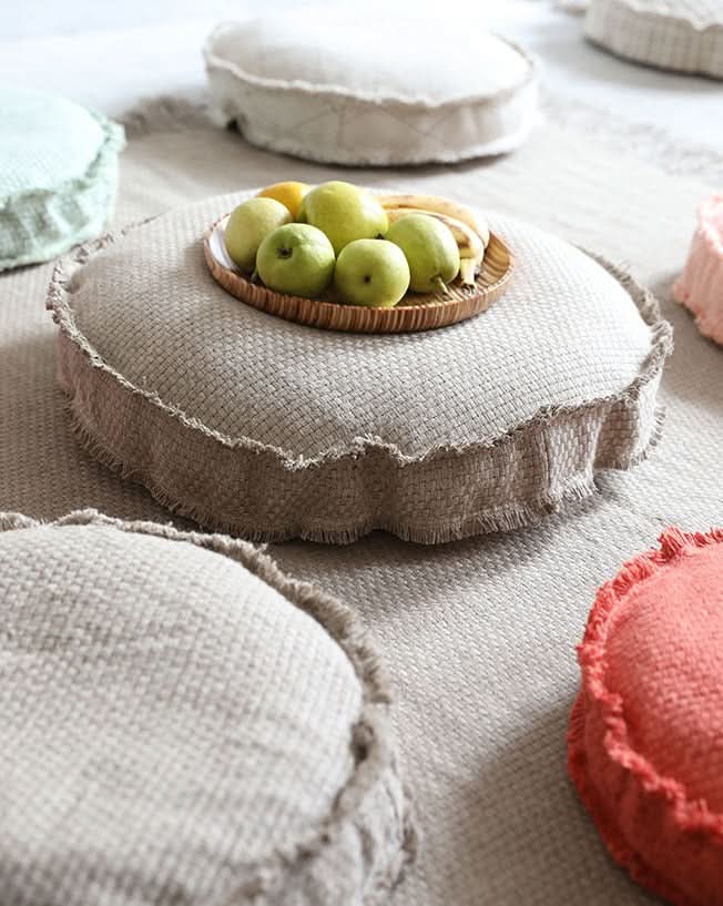 Textured floor cushion in neutral tones with a fringed edge, holding fruit.