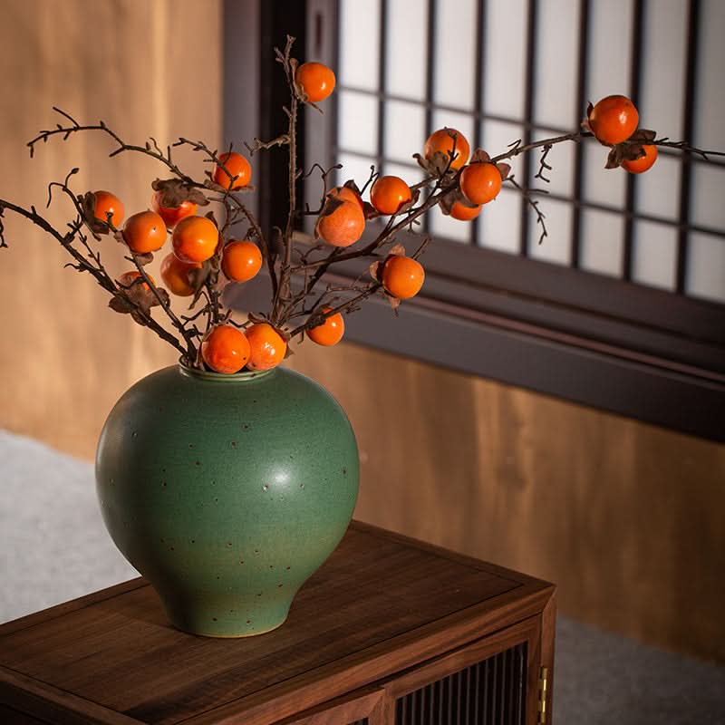 Sage green round vase with artificial persimmon branches on a wooden table.