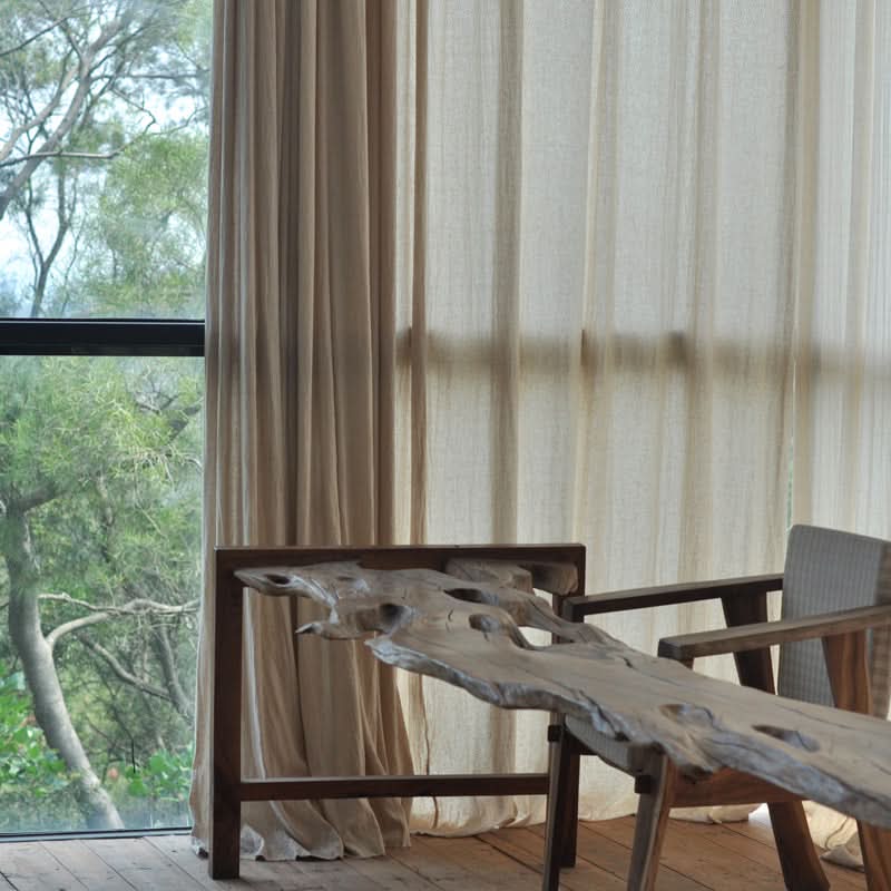 Cream linen curtains hang next to a window, providing a soft backdrop for a rustic wooden table and chair.