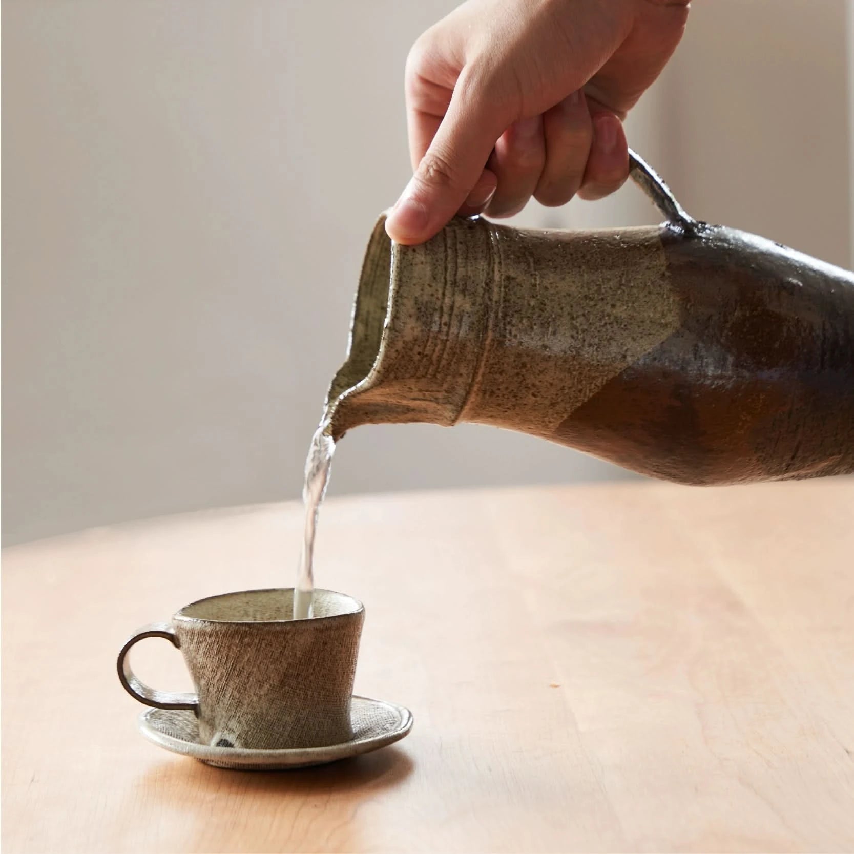 Pouring water from a rough pottery glazed ceramic jug into a matching cup and saucer. Home Decor.
