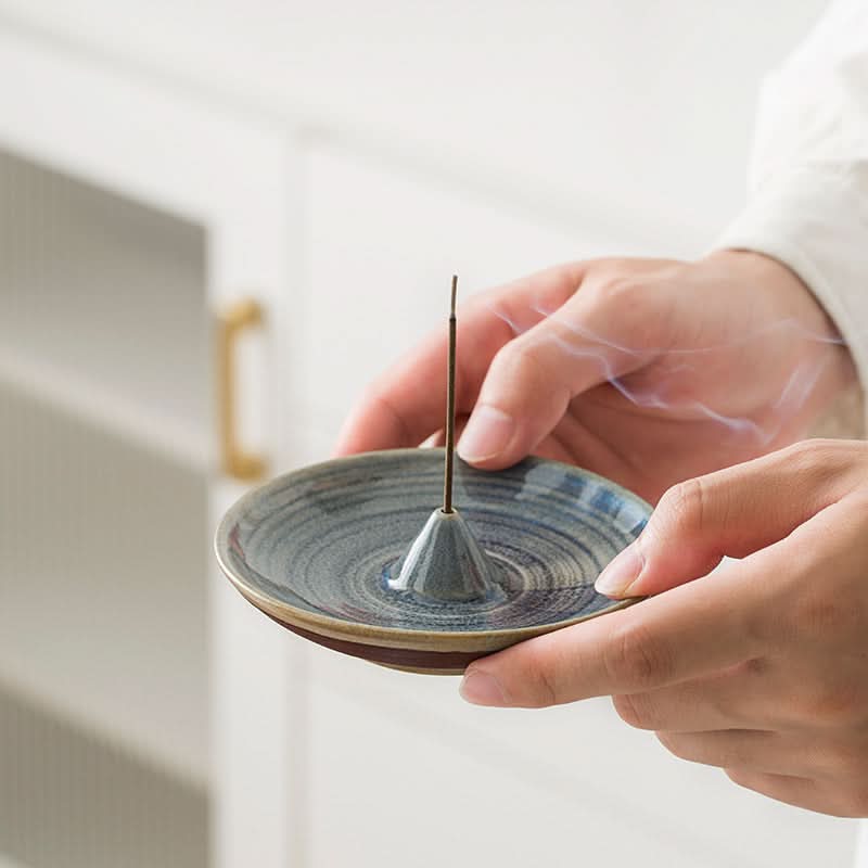 Blue glazed ceramic incense holder with lit incense stick, held in hands.