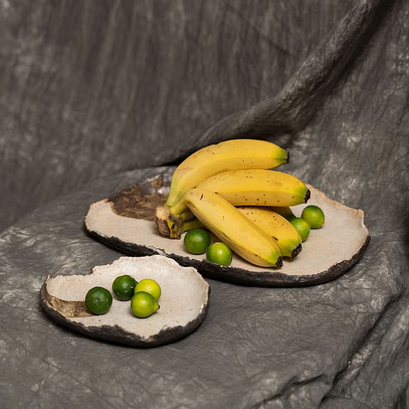 Wabi-sabi irregular beige serving tray with bananas and limes on textured grey fabric.