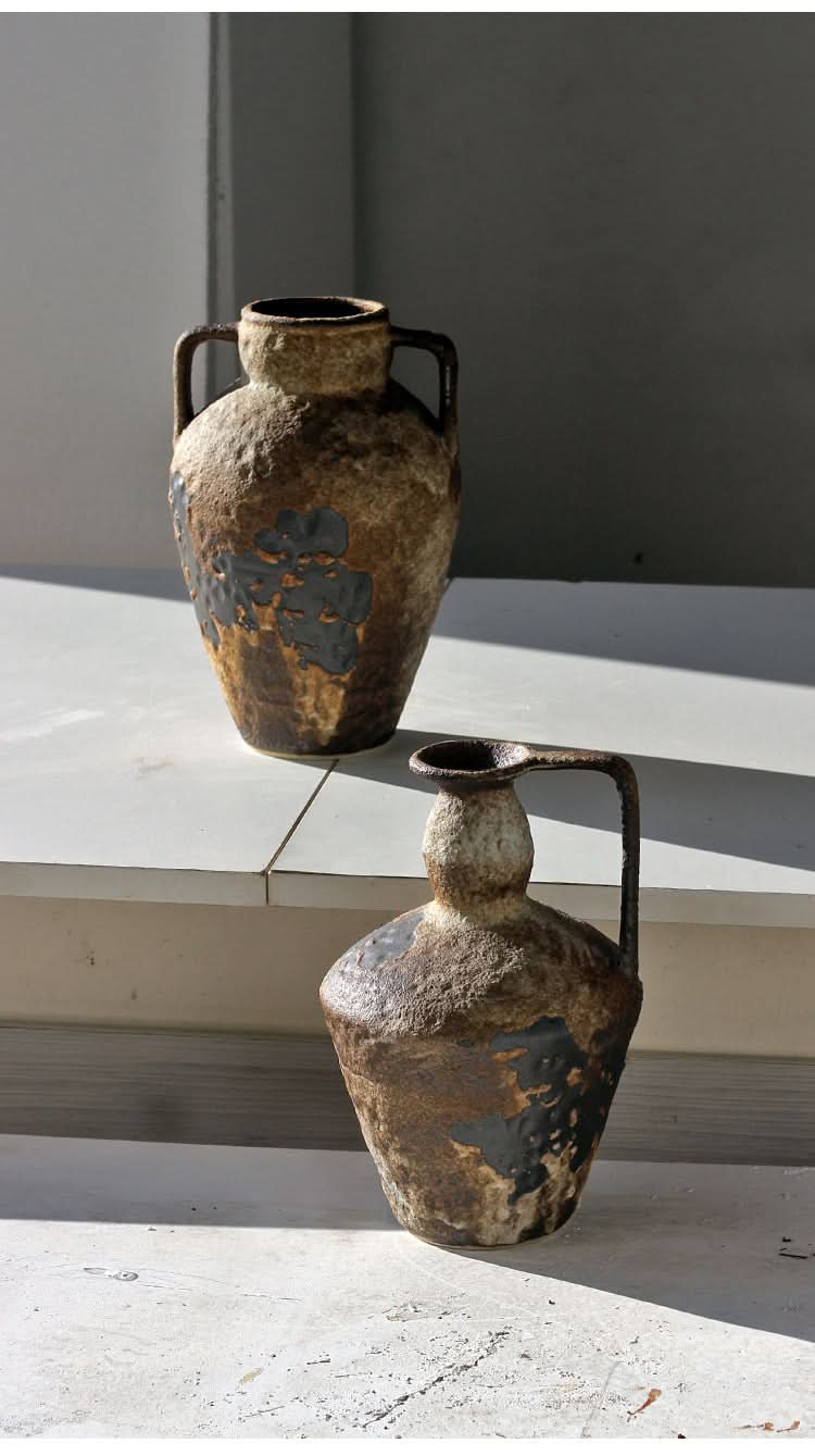 Two rustic, textured small ceramic vases in brown and black, with handles, on a textured surface.
