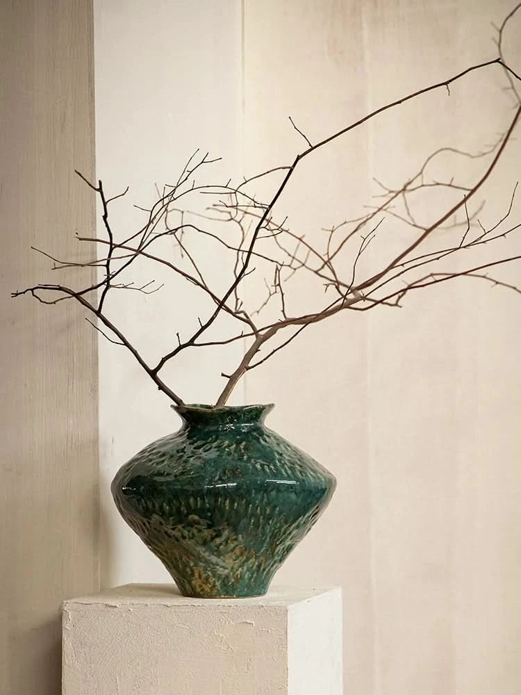 Wabi-Sabi Distressed Rustic Vase with dark green glaze and bare branches on a white pedestal.