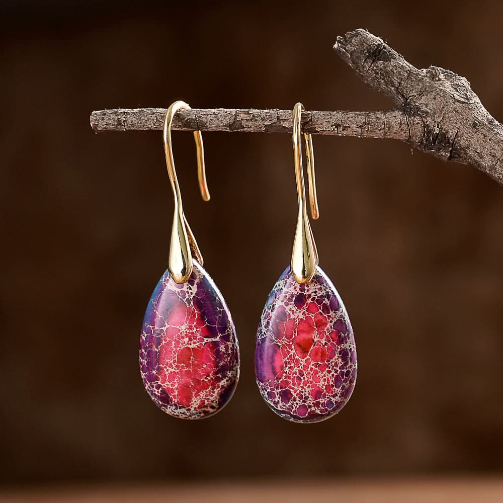 Gold teardrop stone earrings with purple and red jasper cabochons, dangling from a branch.