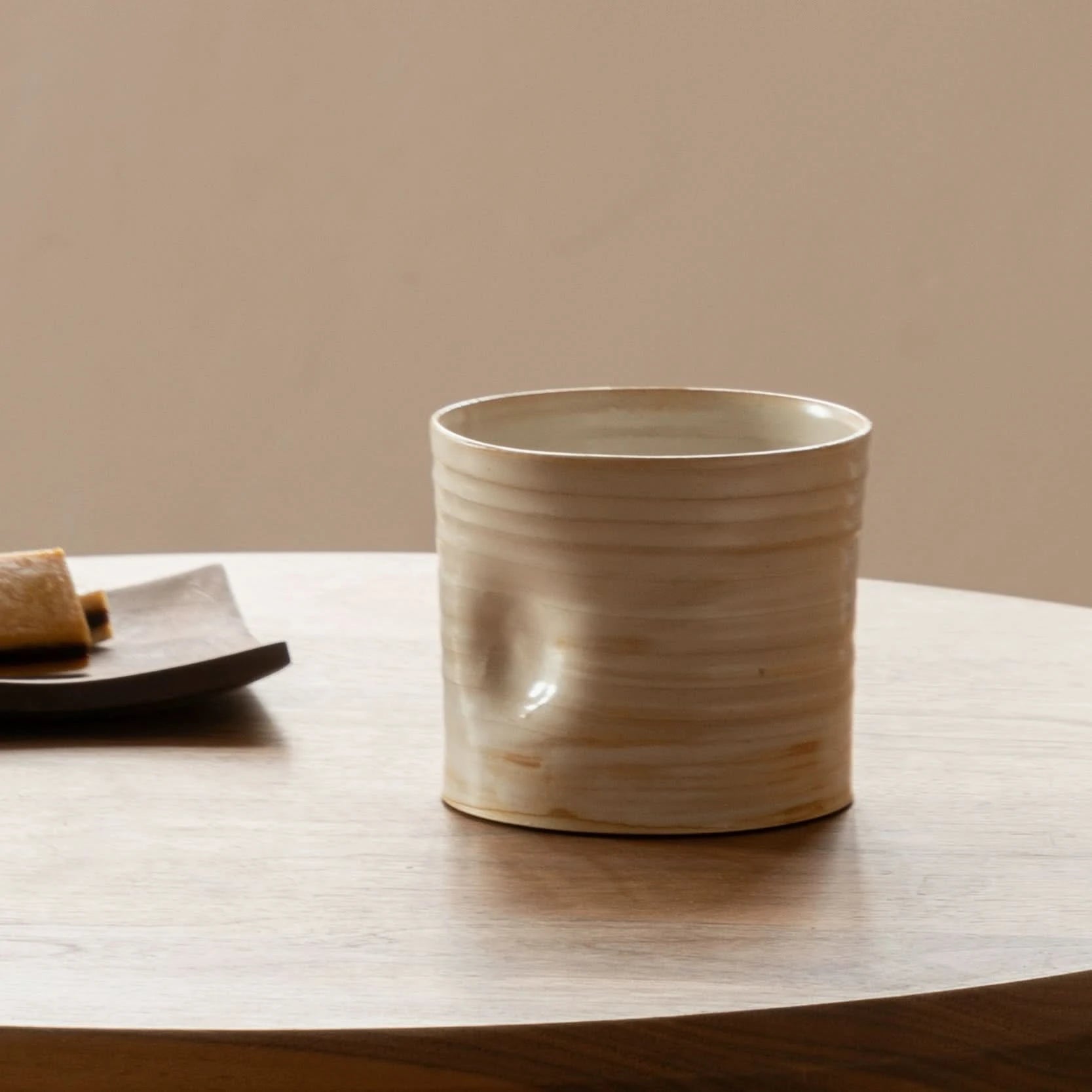 Handmade ceramic dented coffee cup, light beige, on wood table with pastry.