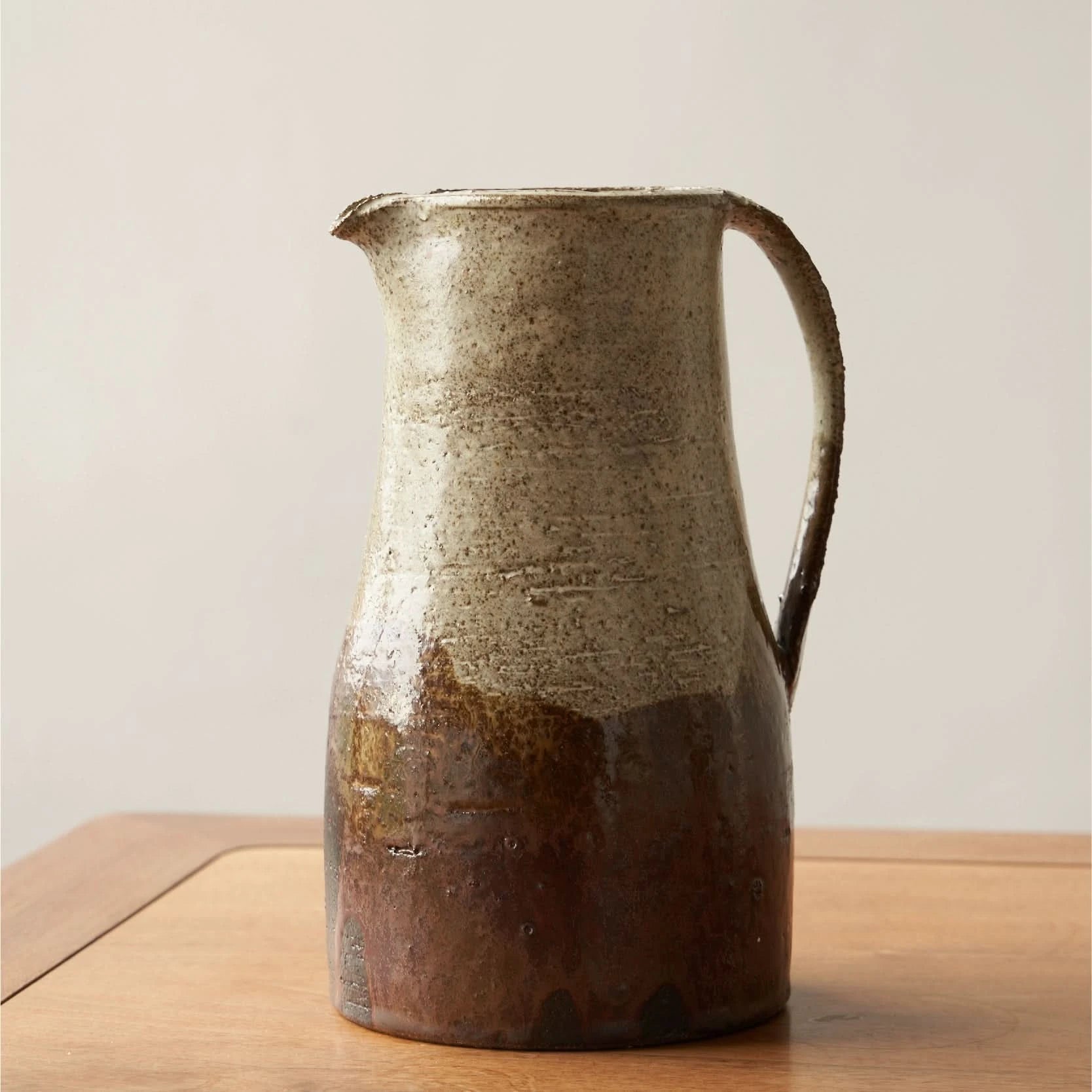 Rough pottery glaze ceramic jug with earthy brown and cream tones, on a wooden surface.