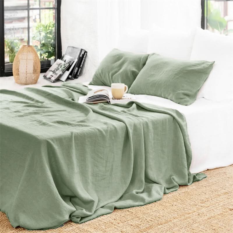 Sage green linen bed sheets with white pillows on a bed, a book and mug on top.