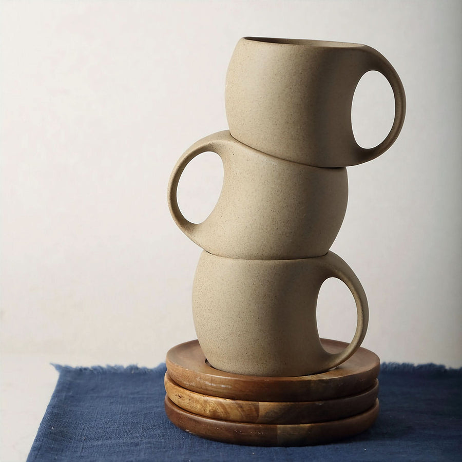 Three speckled beige abstract ceramic mugs with arched handles stacked on wooden coasters on a blue cloth.