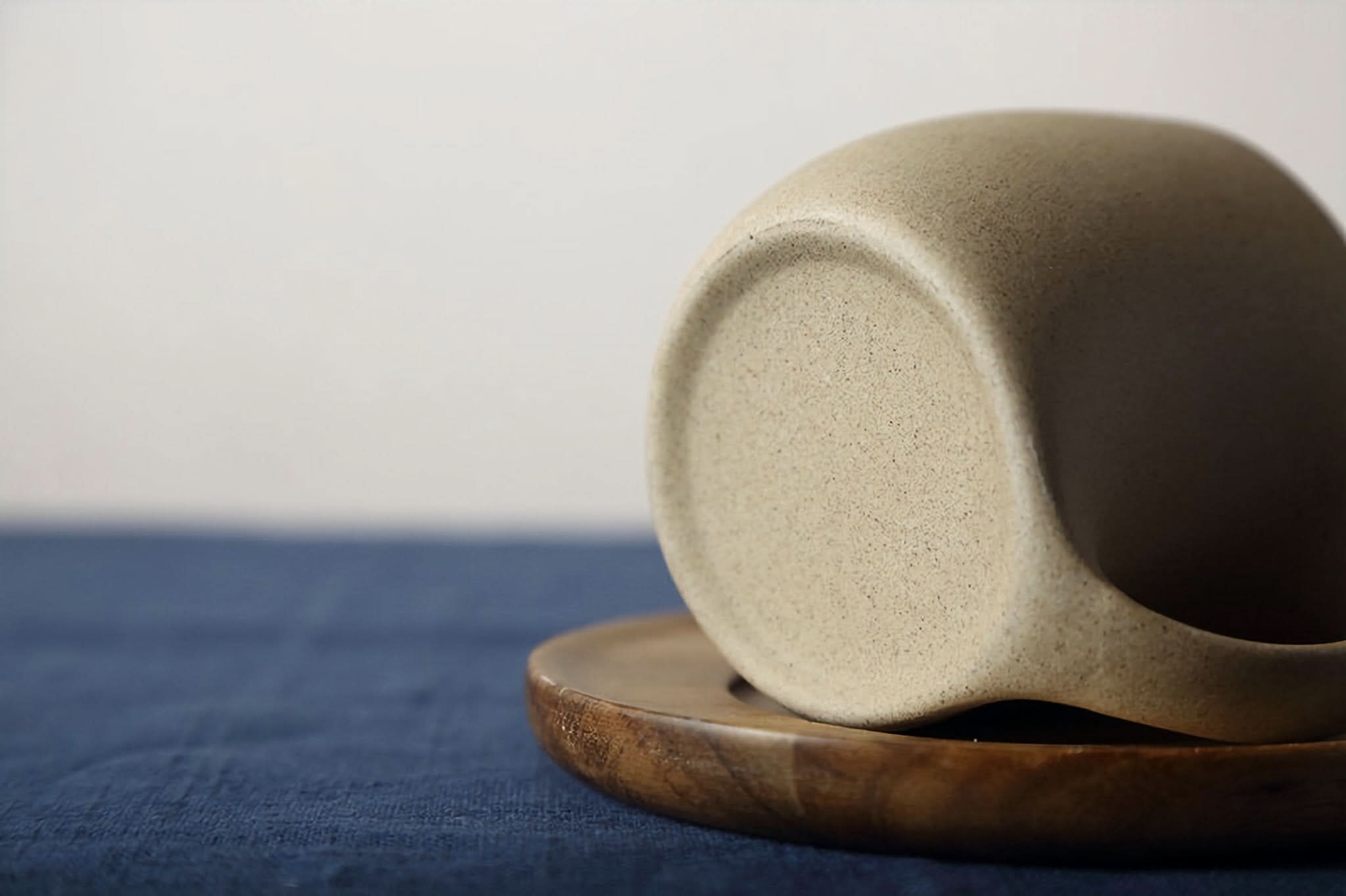 Abstract Ceramic Mug with Arched Handle, speckled beige, on wooden coaster & blue fabric.