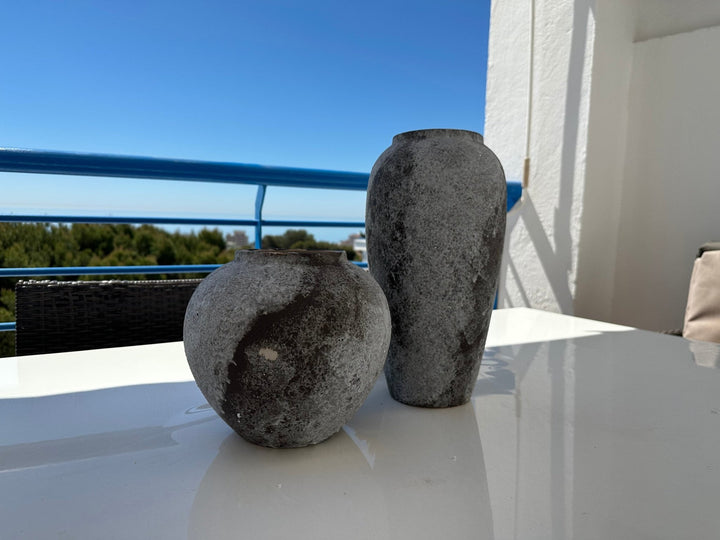 Two textured ceramic vases, dark gray with light gray speckles, sit on a white table outdoors.