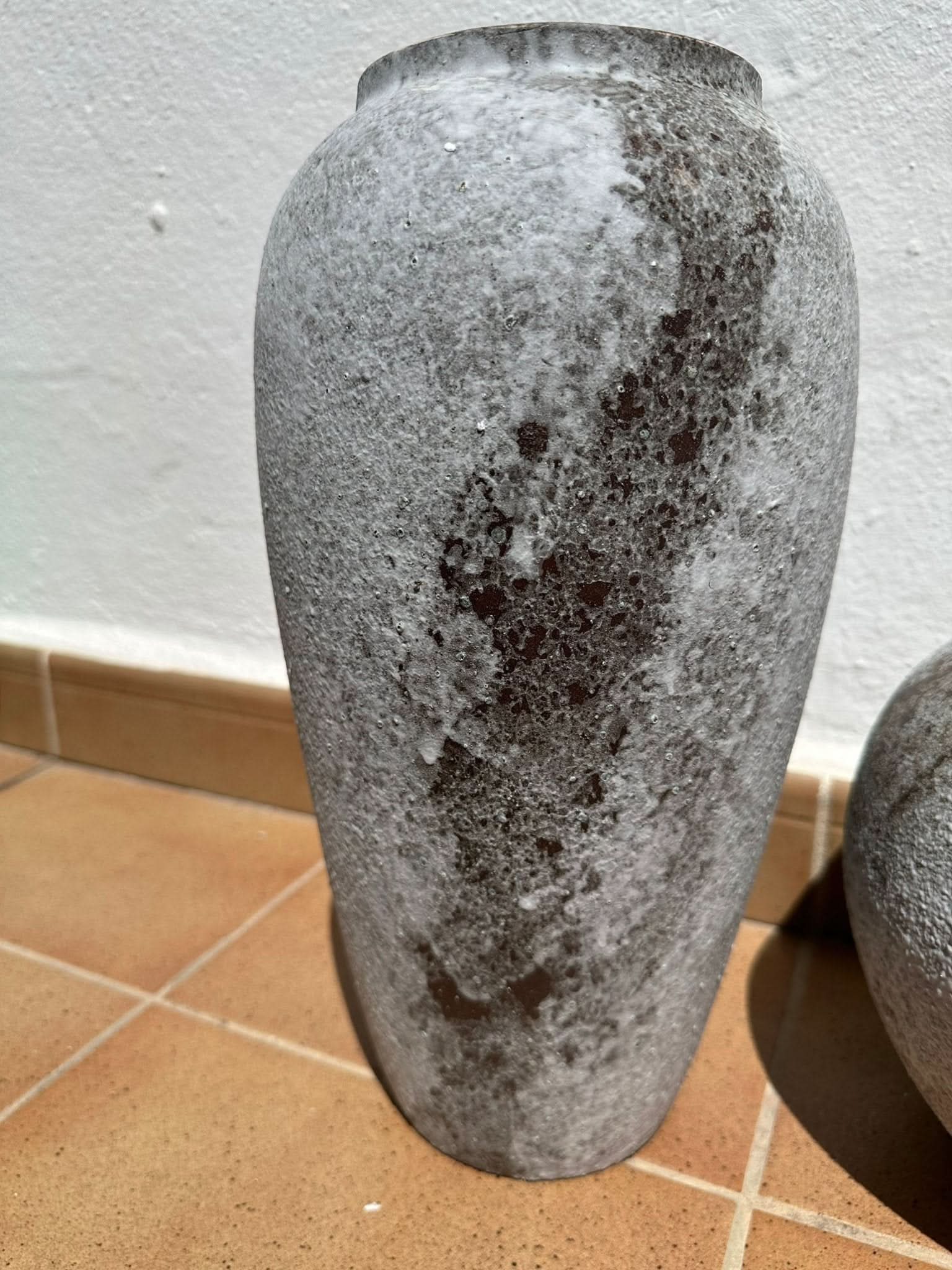 Textured antique effect ceramic vase in grey and dark brown hues, on terracotta tiles.