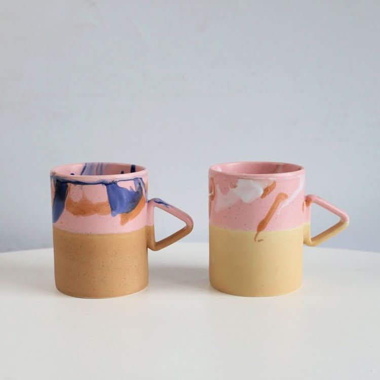 Two Artistic Triangle Handle Stoneware Mugs, pink and yellow with abstract glaze, on white surface.