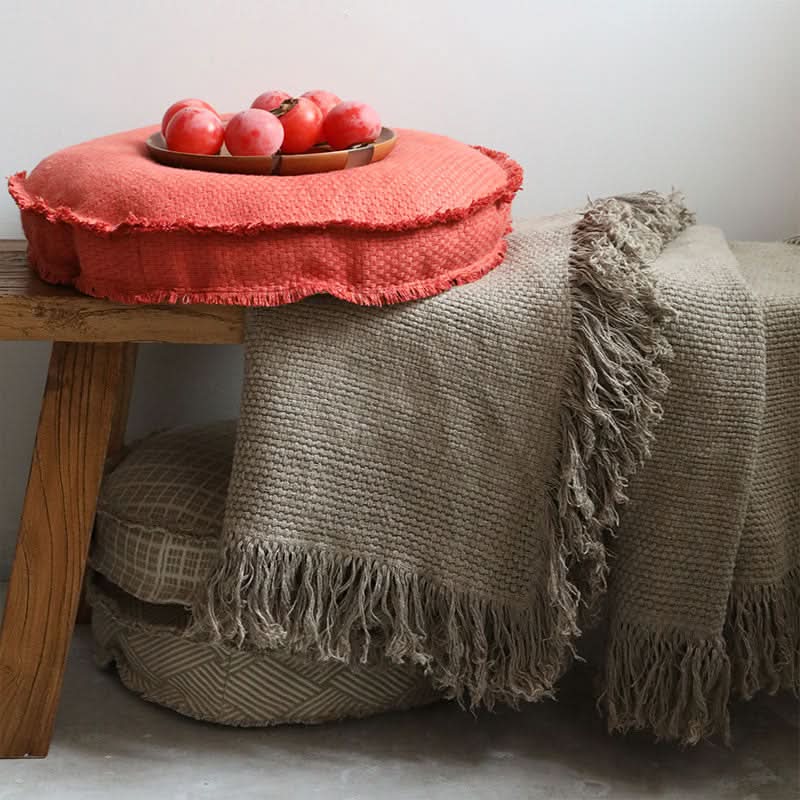 Coral textured floor cushion with fringe on rustic bench beside neutral throw blanket.