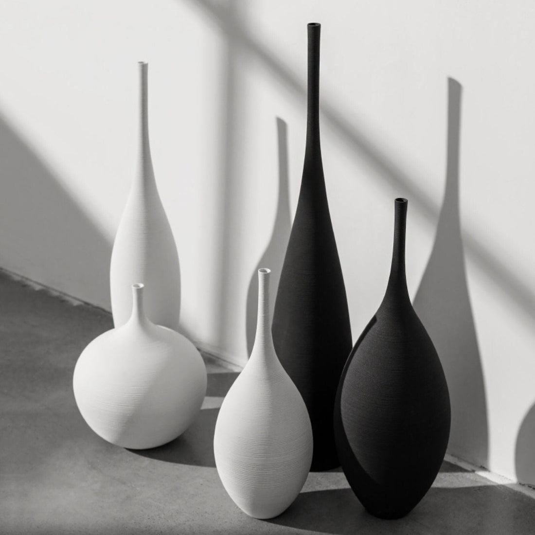 Black and white ceramic vases, various shapes and sizes, on a light gray concrete floor against a white wall.