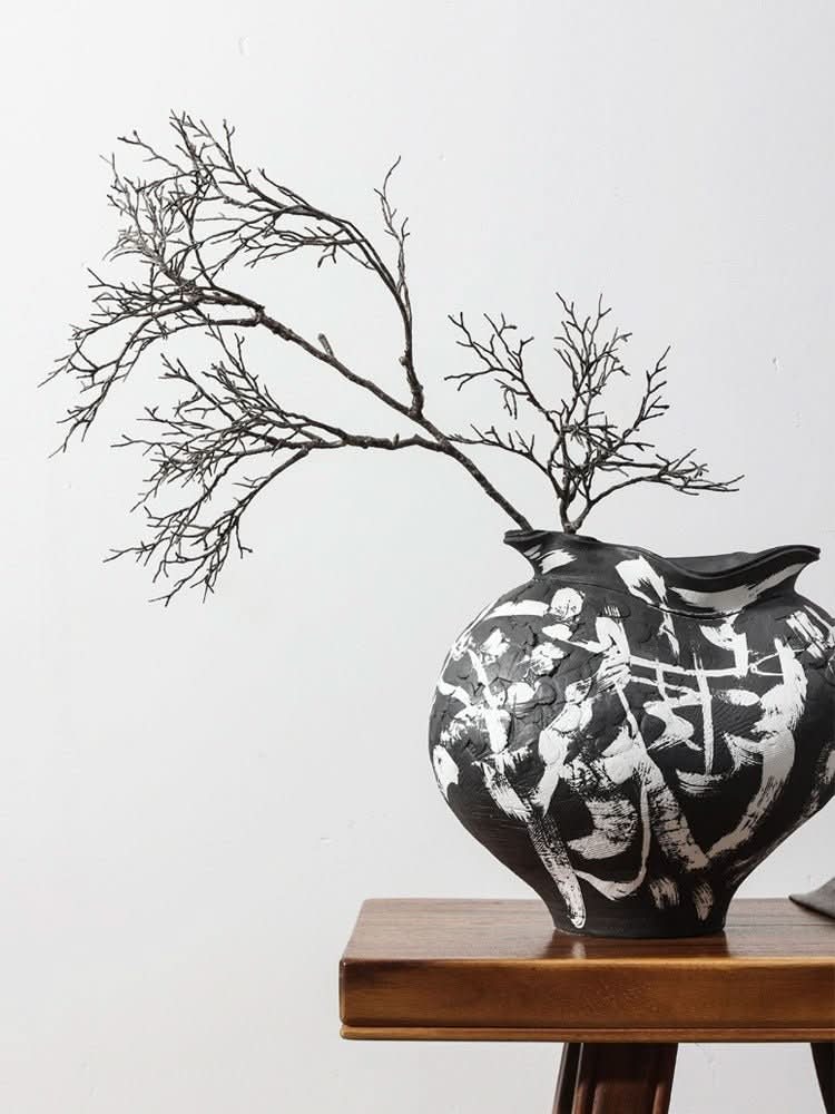 Black irregular wabi-sabi ceramic vase with white brushed effects, holding dried branches on wooden table.
