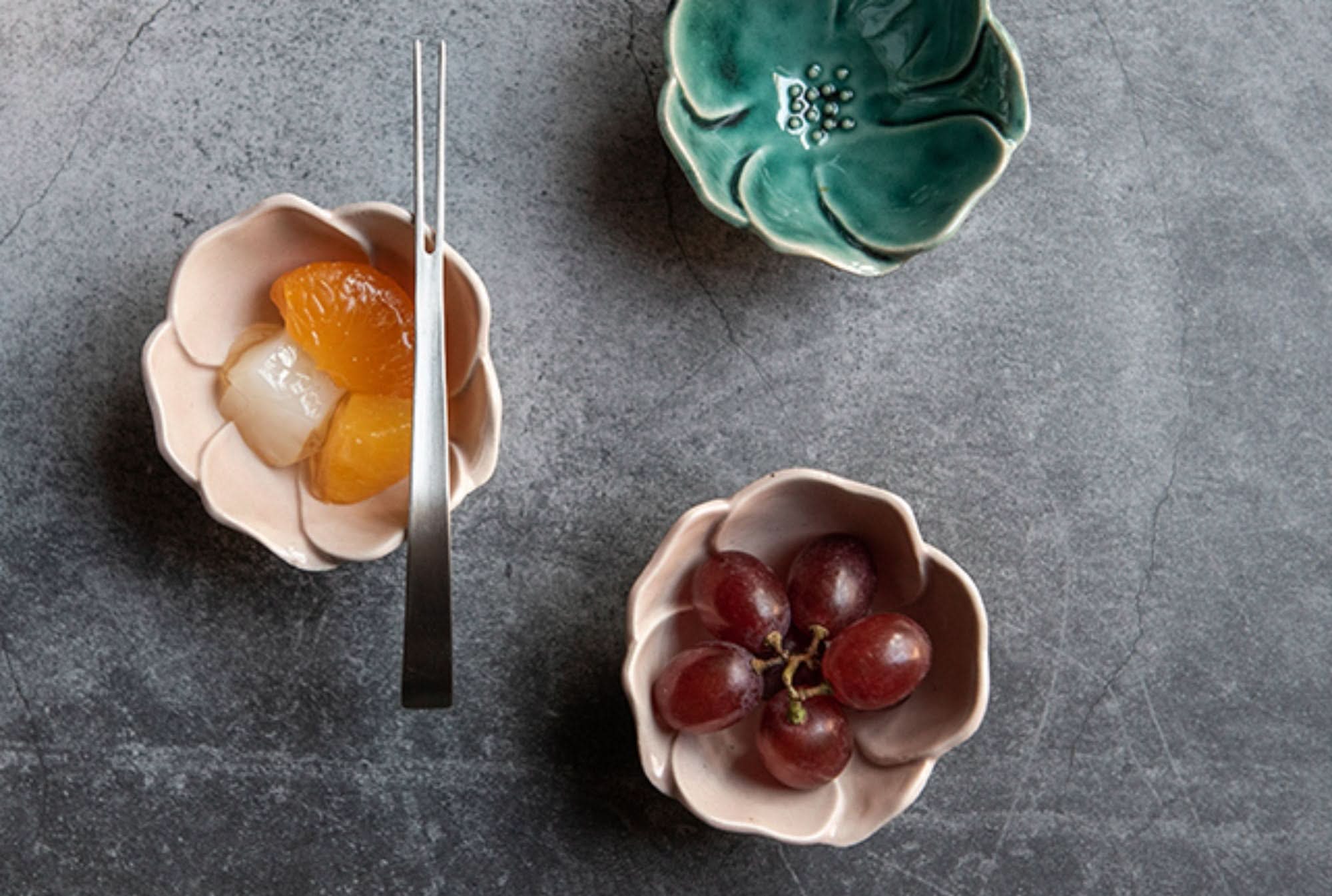 Camellia Ceramic Sauce Bowl: Pink, teal stoneware dishes with fruit, on dark textured surface.