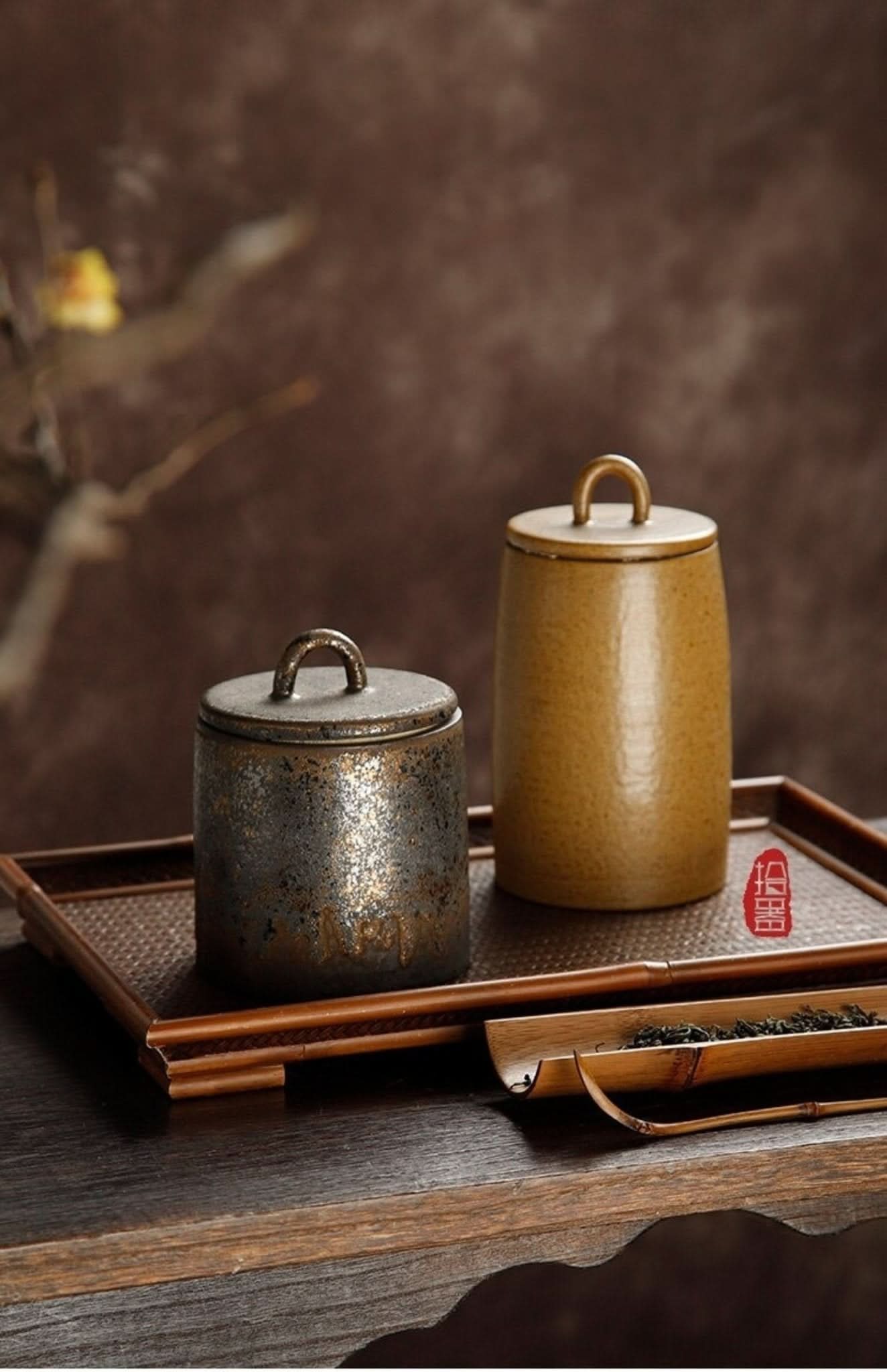 Two ceramic tea canisters, one speckled dark gray, one golden brown, with handles on a bamboo tray.