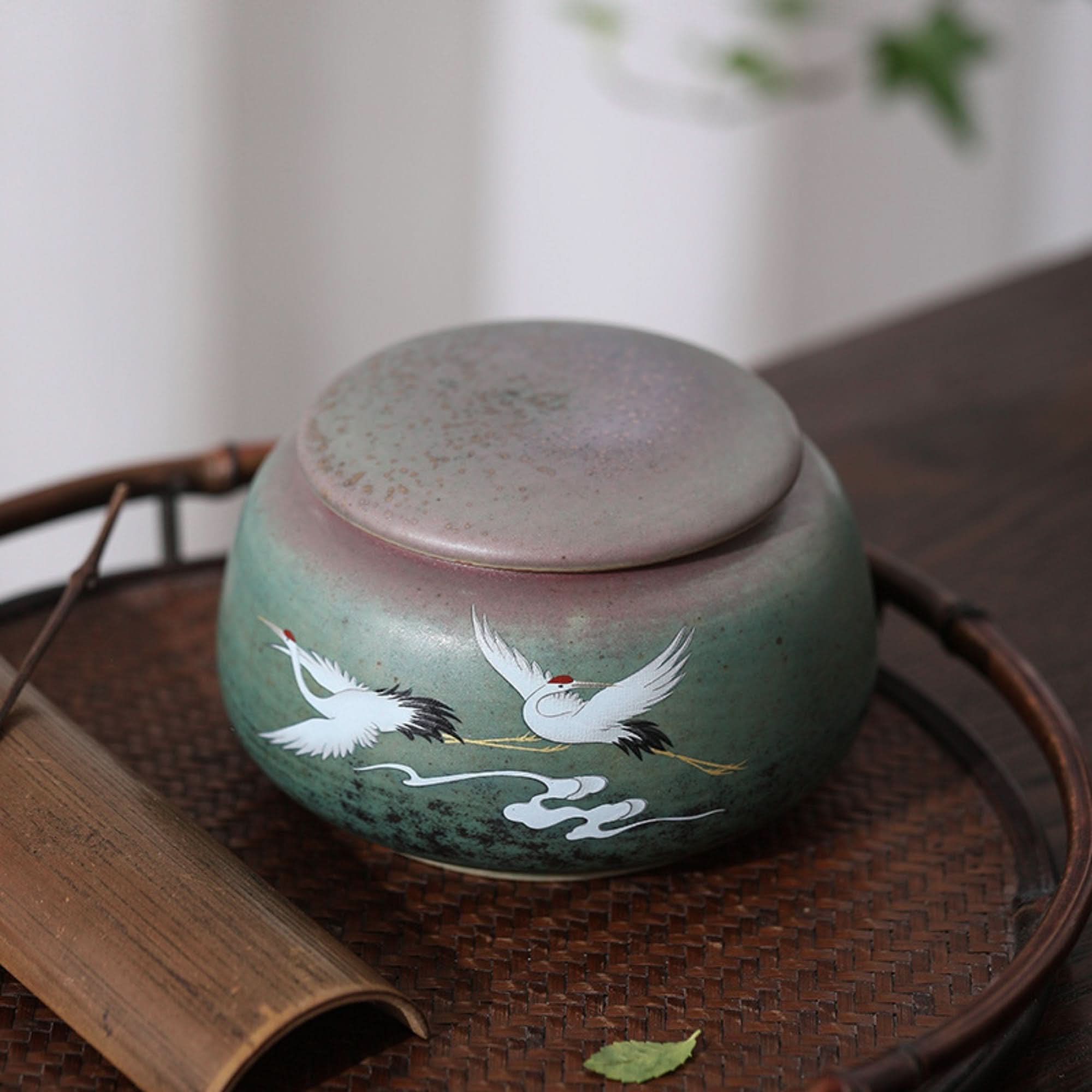 Ceramic tea can with Japanese art illustration, green & pink glaze, on woven tray.