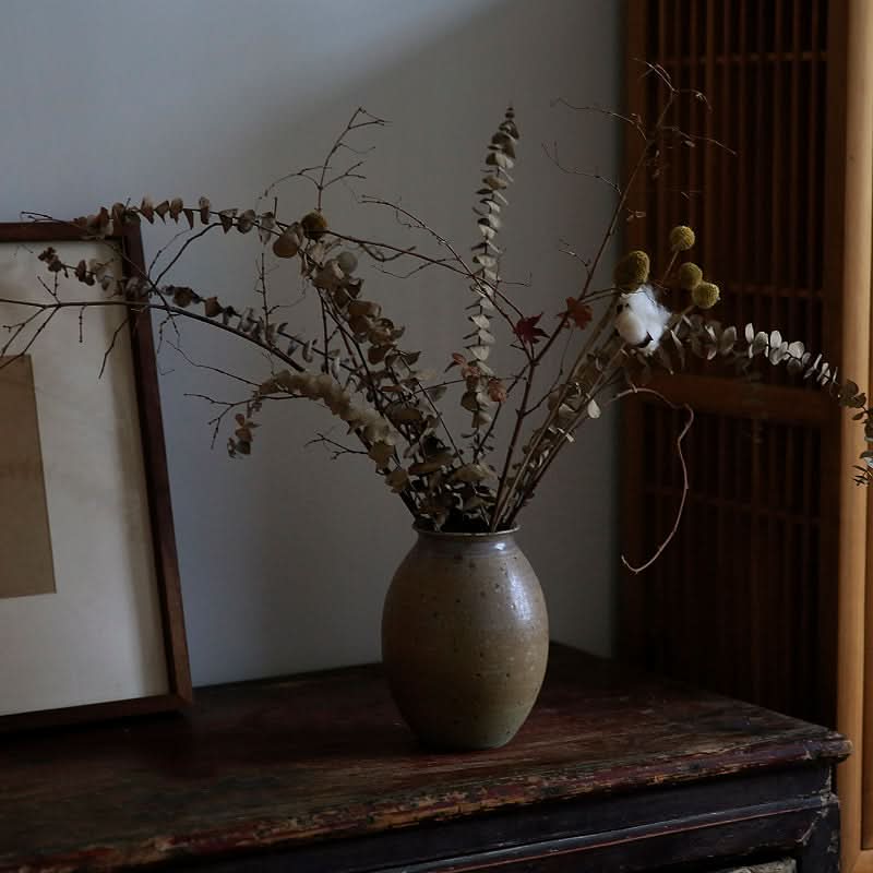 Rustic speckled ceramic vase with dried eucalyptus branches on a wooden dresser.