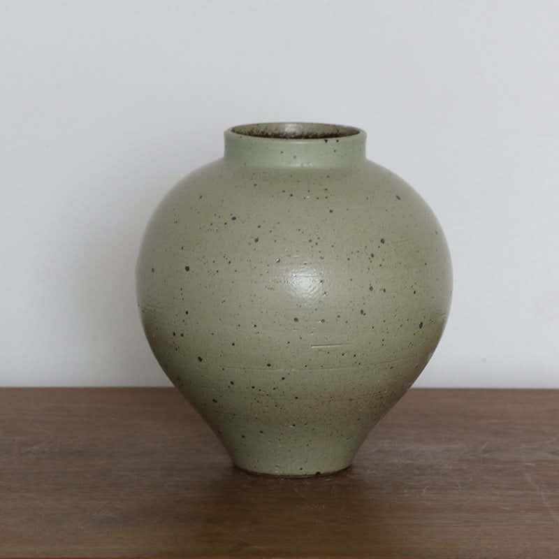 Handmade coarse pottery large round vase in sage green with black speckles on a wooden surface.